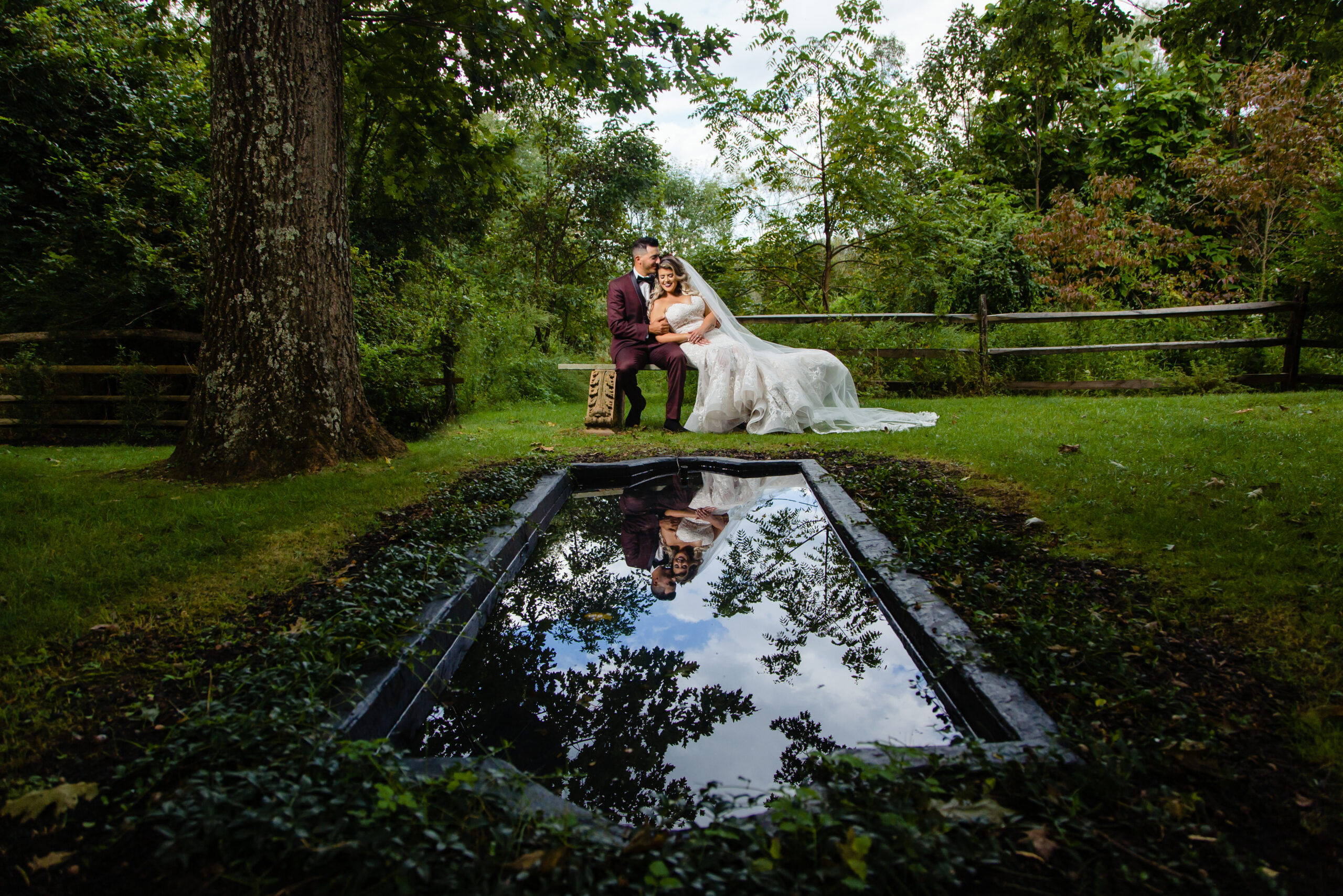 A deeply romantic moment between a newlywed couple, captured at Crossed Keys Estate. They sit on a bench beside a garden pool, their reflections shimmering in the water, creating a poetic and serene scene on their beautiful wedding day. This touching moment was skillfully captured by North Jersey wedding photographer Jarot Bocanegra