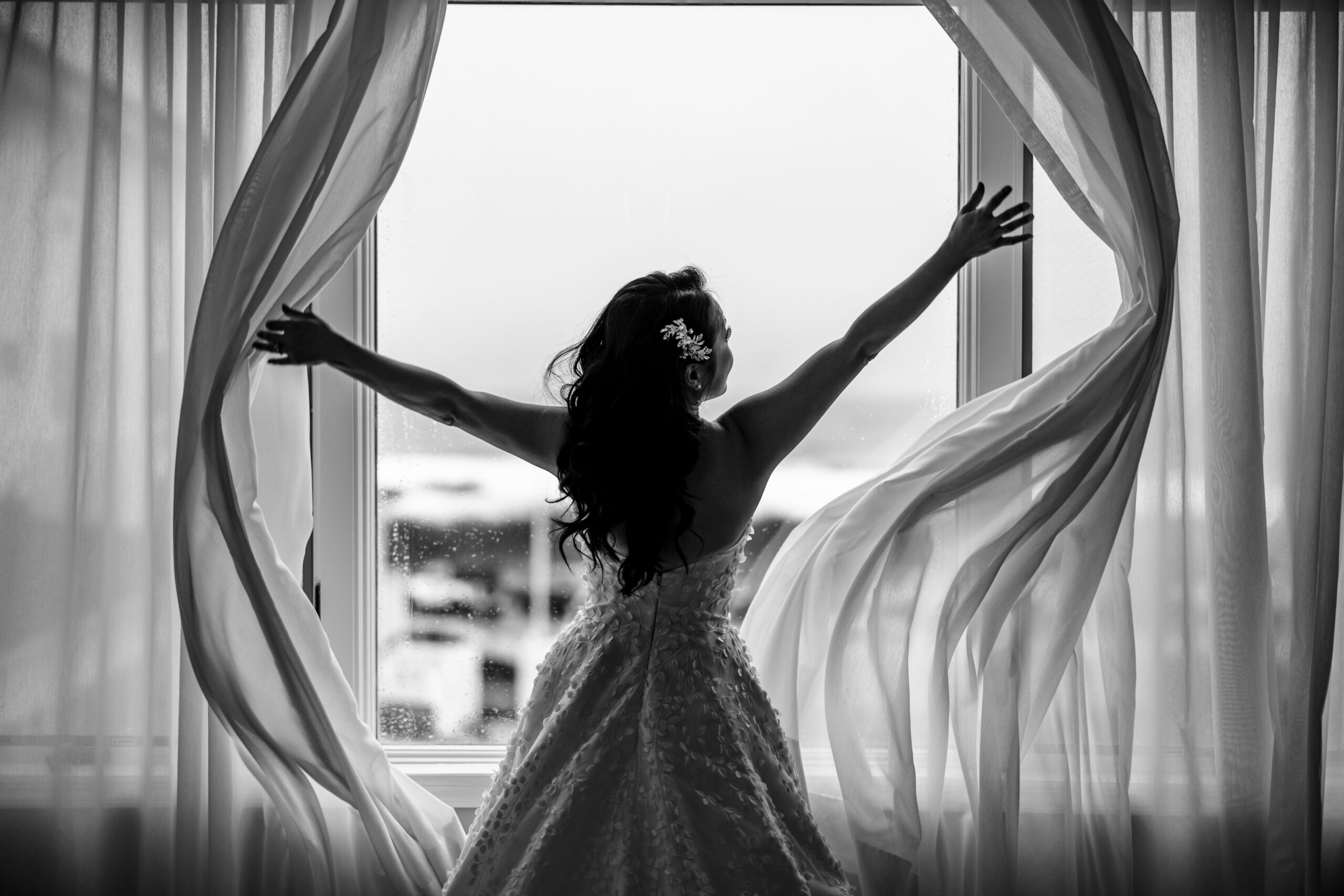 Graceful bride portrait as she opens the curtains on her wedding day, welcoming the light of a new beginning. This elegant moment was skillfully captured by North Jersey wedding photographer Jarot Bocanegra