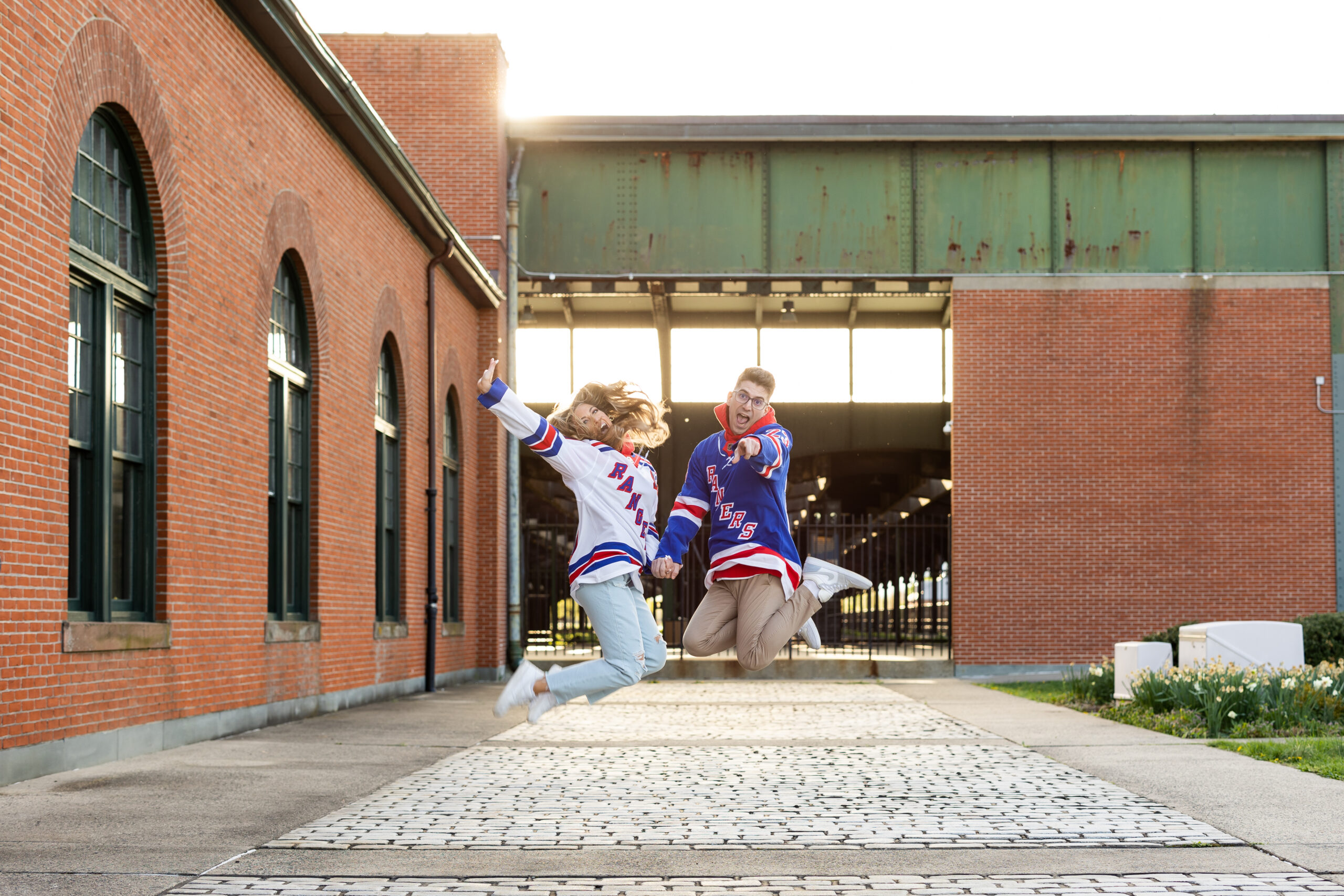 A couple captured by New Jersey Wedding Photographer Jarot Bocanegra, jumping in the air in front of a brick building.