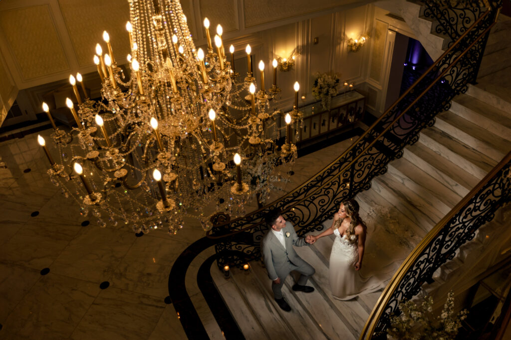 A bride and groom, captured by New Jersey Wedding Photographer Jarot Bocanegra, standing on a staircase with a chandelier.