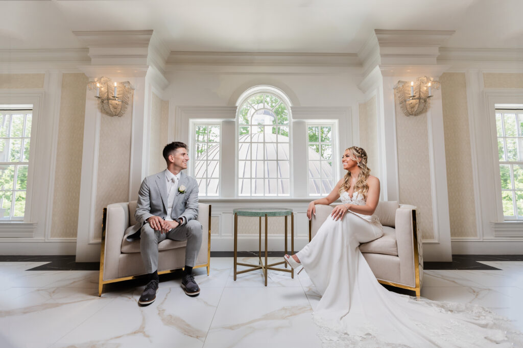 A bride and groom, captured by New Jersey Wedding Photographer Jarot Bocanegra, sitting in chairs in a room.