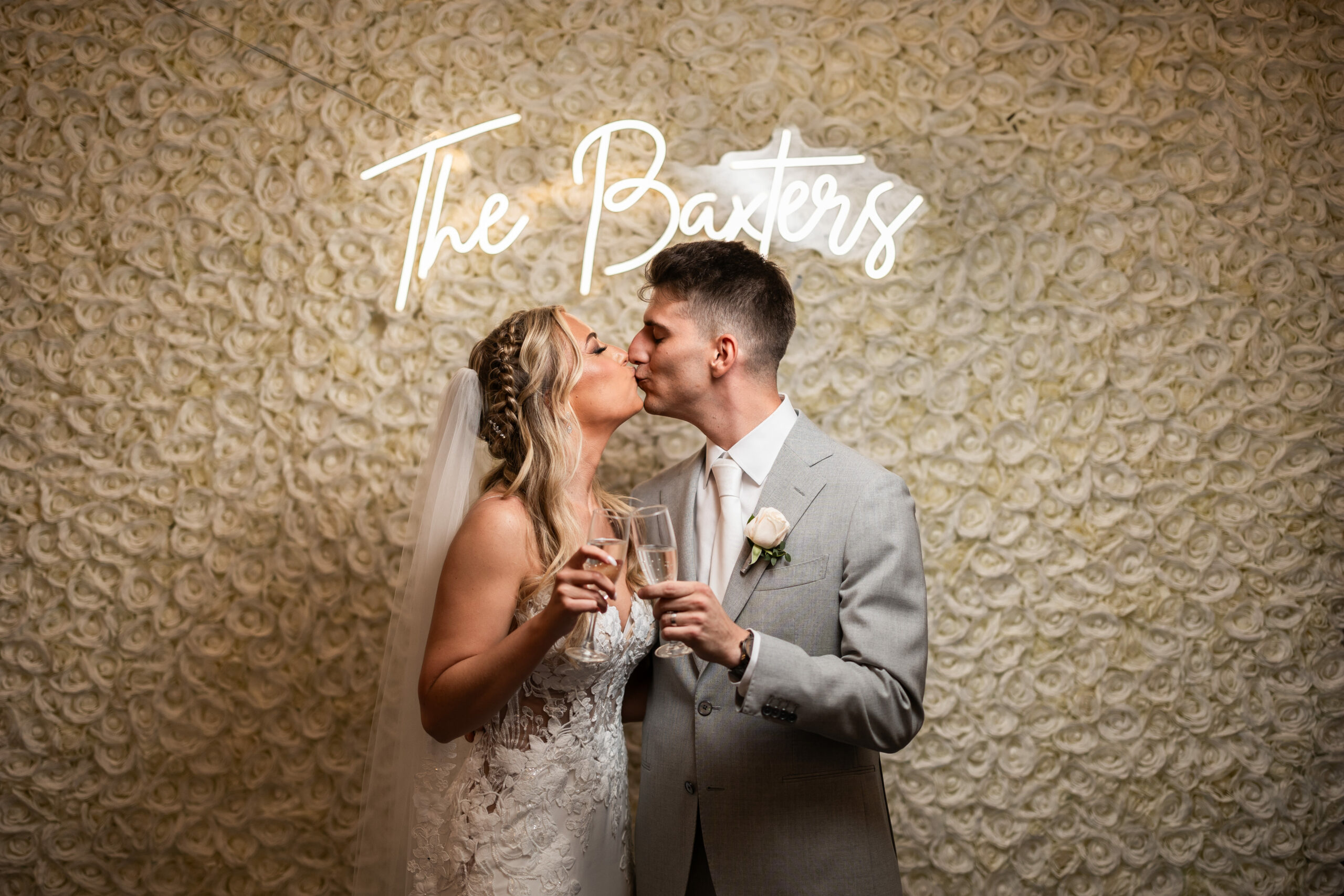 A loving couple shares a kiss after their ceremony, standing before a stunning flower wall with their names displayed in a neon sign. This heartwarming moment was beautifully captured by Jarot Bocanegra Photography
