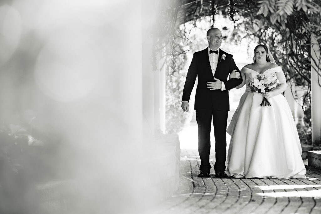 Captured by New Jersey Wedding Photographer Jarot Bocanegra, a black and white photo captures the bride and groom walking down a pathway.
