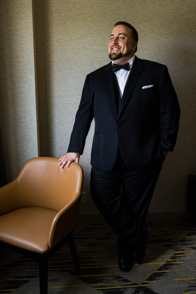 A man in a tuxedo leaning against a chair, elegantly captured by New Jersey Wedding Photographer Jarot Bocanegra.