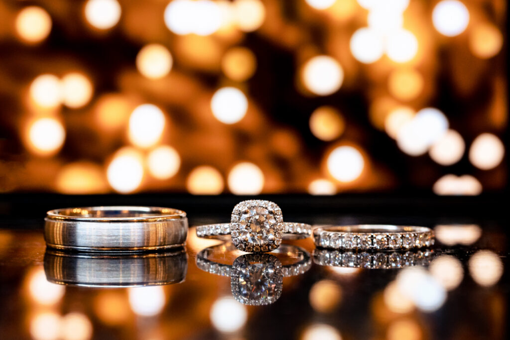 Wedding rings captured by New Jersey Wedding Photographer Jarot Bocanegra, beautifully displayed on a table with enchanting lights behind them.