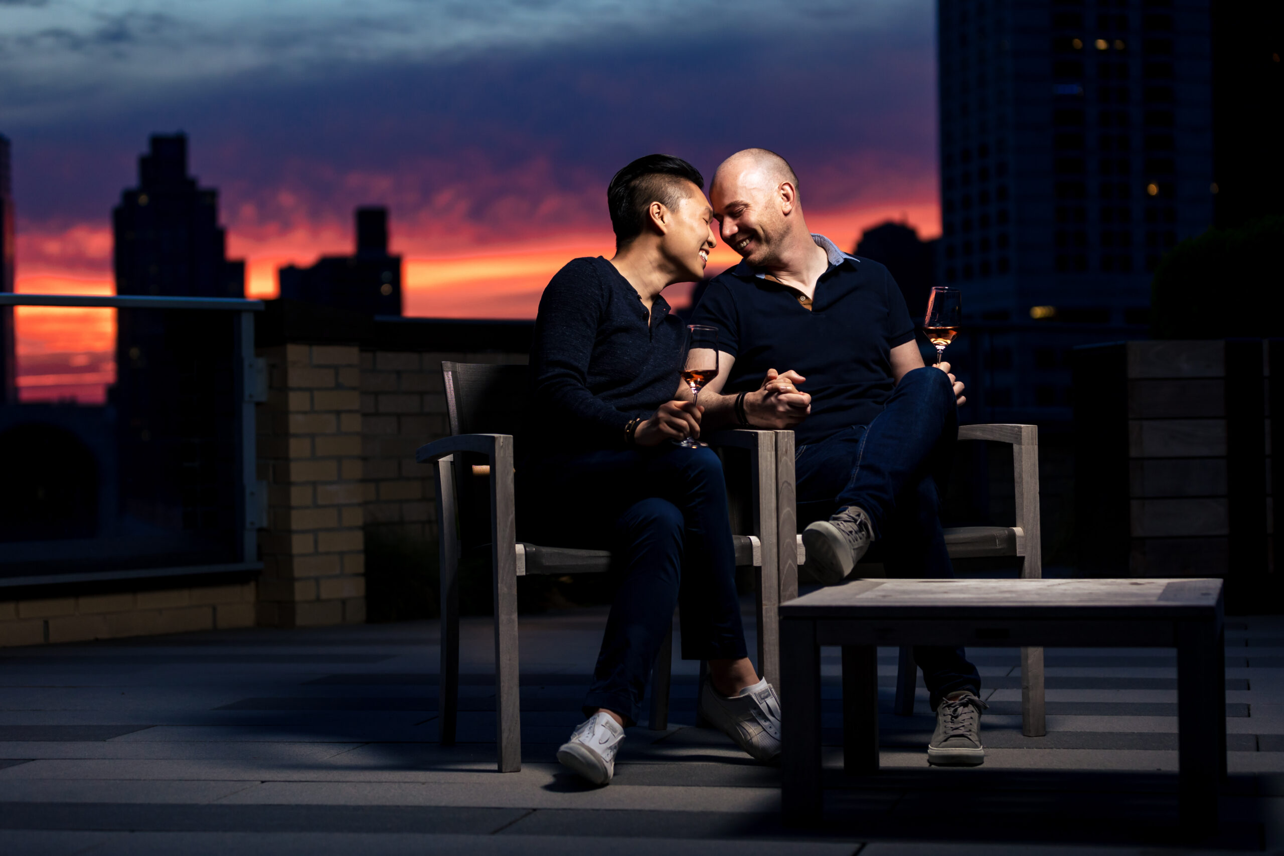 Engagement session portrait of a joyful LGBTQ+ couple on a New York City rooftop, with a breathtaking sunset over the city skyline in the background., captured by North Jersey wedding photographer Jarot Bocanegra