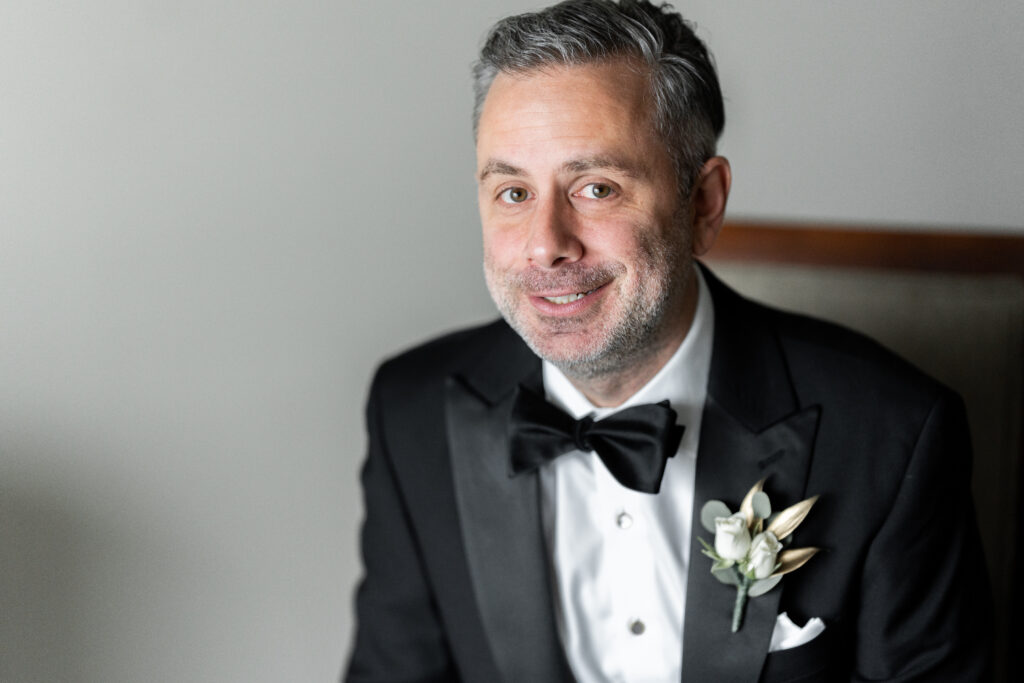 A man in a tuxedo smiles while sitting in a chair, captured by New Jersey Wedding Photographer Jarot Bocanegra.