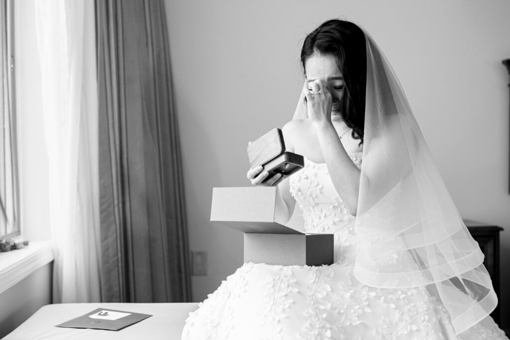 A bride, captured by New Jersey Wedding Photographer Jarot Bocanegra, is sitting on a bed holding a box.