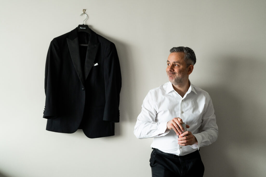 A man captured by New Jersey Wedding Photographer Jarot Bocanegra standing next to a tuxedo hanging on a wall.