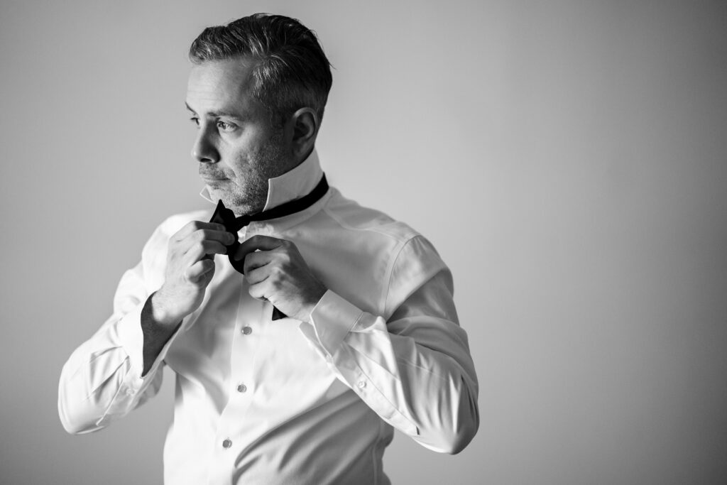 Captured by New Jersey Wedding Photographer Jarot Bocanegra, a black and white photo showcases a man adjusting his bow tie.