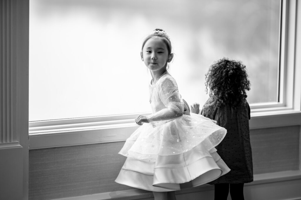 Two little girls in white dresses standing by a window, captured by New Jersey Wedding Photographer Jarot Bocanegra.