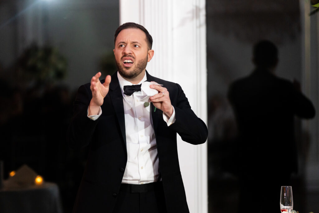 A man captured by New Jersey Wedding Photographer Jarot Bocanegra, in a tuxedo, speaking into a microphone.