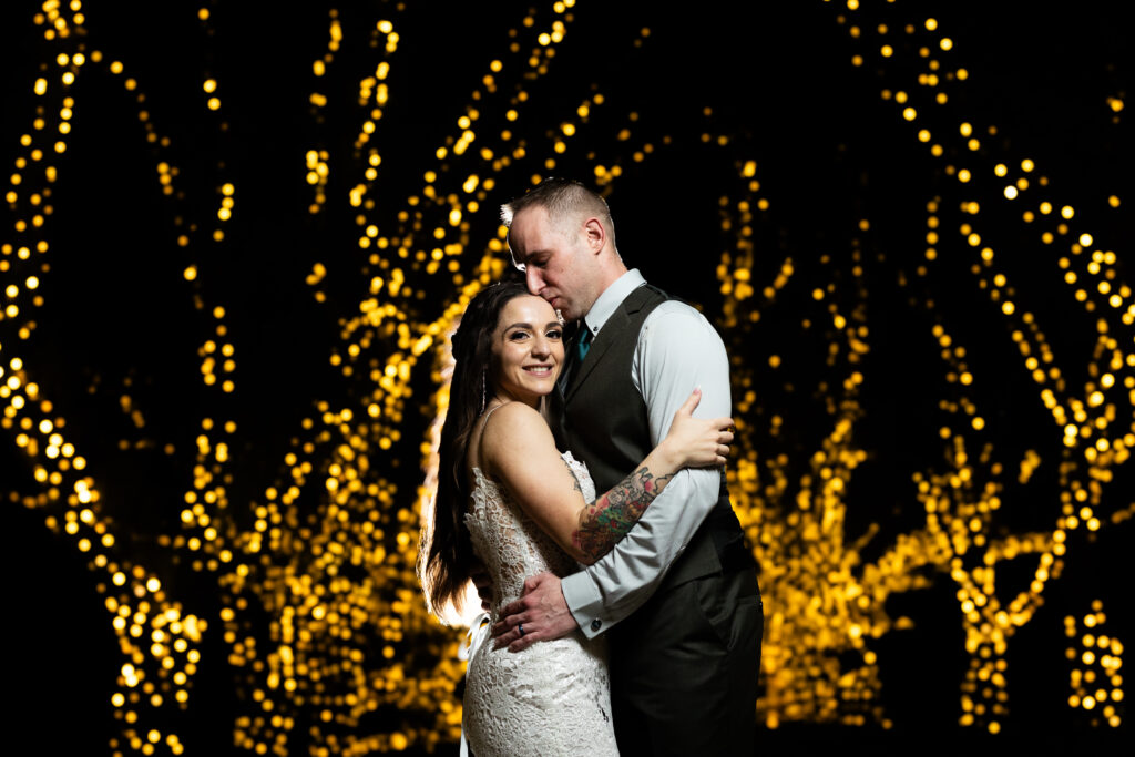 A bride and groom embrace in front of a lit up tree, captured by New Jersey Wedding Photographer.