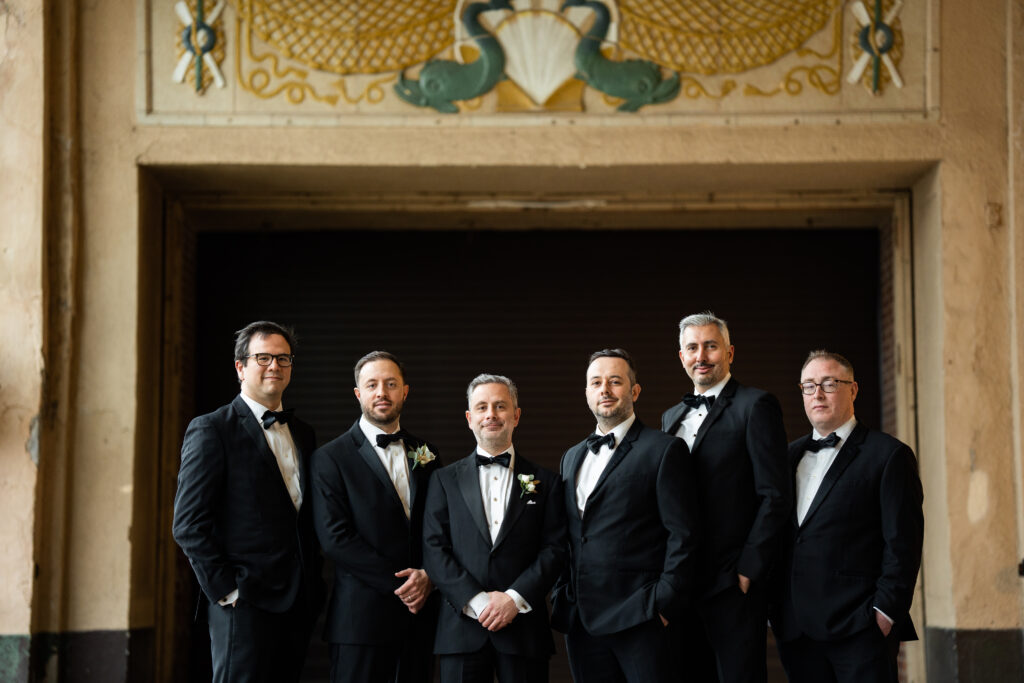 A group of groomsmen in tuxedos standing in front of an ornate door, captured by New Jersey Wedding Photographer Jarot Bocanegra.