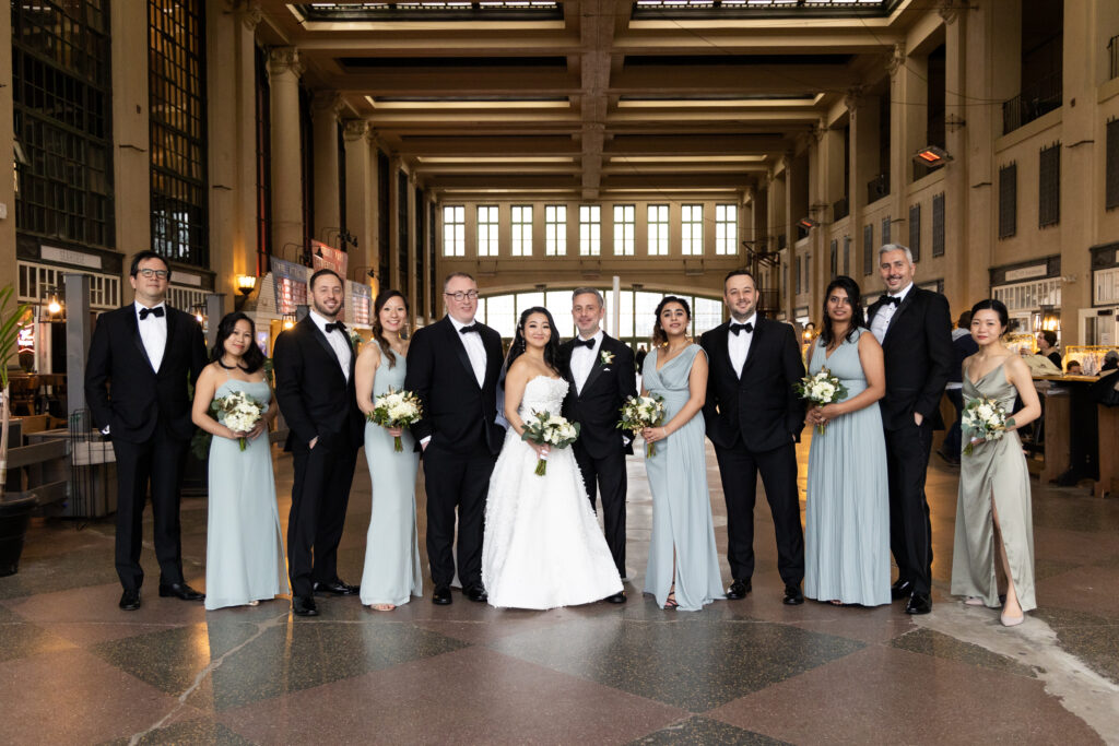 A group of bridesmaids and groomsmen pose for a photo in a large building, captured by New Jersey Wedding Photographer Jarot Bocanegra.