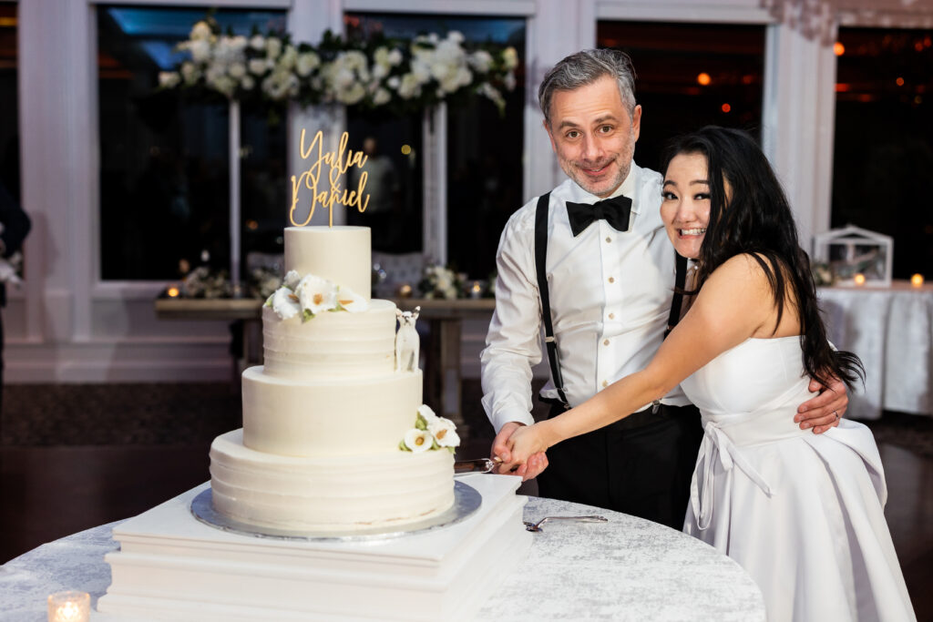 A bride and groom cutting into a white wedding cake at The Mill Lakeside Manor, captured by New Jersey Wedding Photographer Jarot Bocanegra.